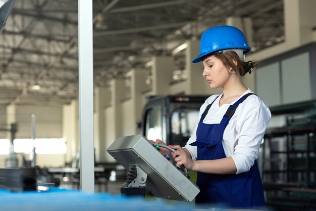 Eine junge attraktive Dame der Vorderansicht im blauen Bauanzug und in den Helm, die Maschinen im Hangar steuern, die während der Architektur des Tagesgebäudes arbeiten