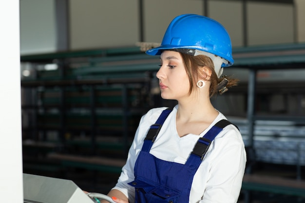 Eine junge attraktive Dame der Vorderansicht im blauen Bauanzug und in den Helm, die Maschinen im Hangar steuern, die während der Architektur des Tagesgebäudes arbeiten