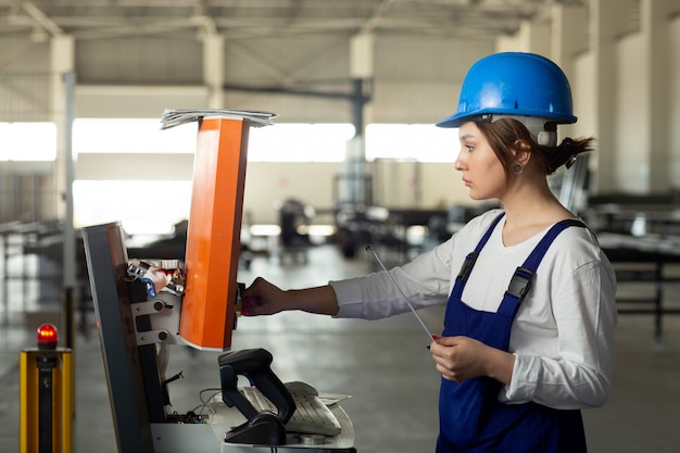 Eine junge attraktive Dame der Vorderansicht im blauen Bauanzug und in den Helm, die Maschinen im Hangar während des Architekturbaus der Tagesgebäude steuern