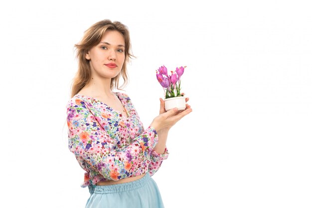 Eine junge attraktive Dame der Vorderansicht im bunten Blumendesignhemd und im blauen Rock, die lila Blumenpflanze auf dem Weiß halten