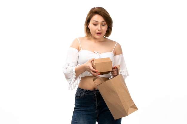 Eine junge attraktive Dame der Vorderansicht im weißen Hemd und in den blauen Jeans, die braunes Paket halten, das auf dem Weiß lächelt