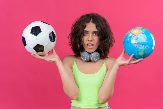 Eine junge attraktive Frau mit kurzen Haaren im grünen Erntedach in Kopfhörern, die Globus und Fußball halten