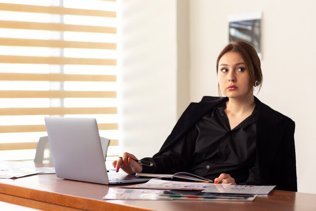 Eine junge attraktive Geschäftsfrau der Vorderansicht in der schwarzen Jacke des schwarzen Hemdes unter Verwendung ihrer silbernen Laptop-Schreiblesung, die in ihrem Büroarbeitsjobgebäude arbeitet