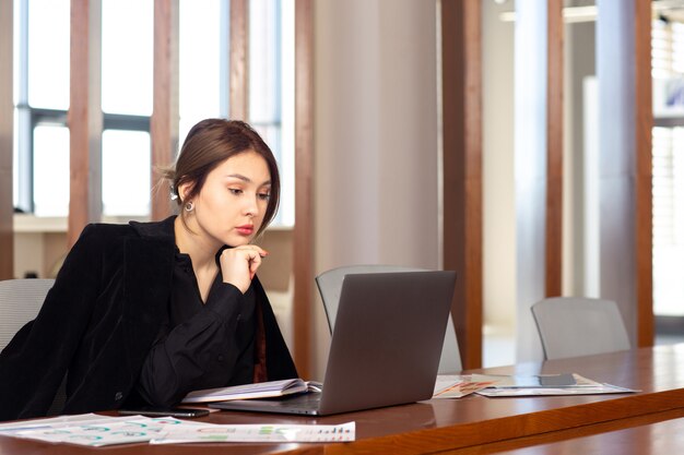 Eine junge attraktive Geschäftsfrau der Vorderansicht in der schwarzen Jacke des schwarzen Hemdes unter Verwendung ihres silbernen Laptops, der innerhalb ihres Büroarbeitsjobgebäudes arbeitet