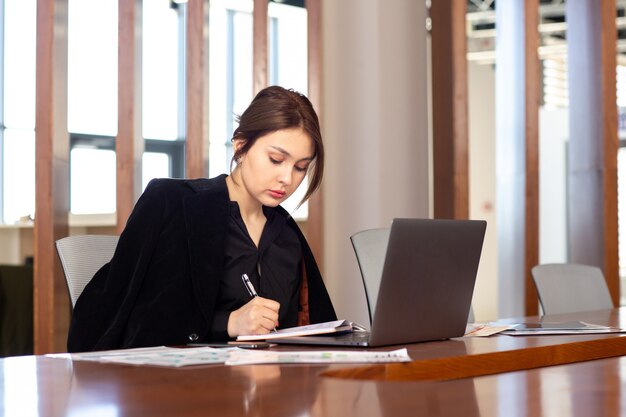 Eine junge attraktive Geschäftsfrau der Vorderansicht in der schwarzen Jacke des schwarzen Hemdes unter Verwendung ihres silbernen Laptops, der Notizen schreibt, die innerhalb ihres Büroarbeitsjobgebäudes arbeiten