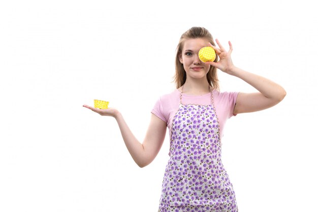 Eine junge attraktive Hausfrau der Vorderansicht im bunten Umhang des rosa Hemdes, das gelbes Ding hält, das auf der weiblichen Küche der weißen Hintergrundküche lächelt