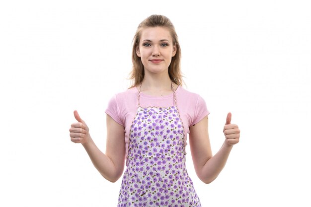 Eine junge attraktive Hausfrau der Vorderansicht im bunten Umhang des rosa Hemdes lächelnd posierend, das fantastische Ausdrucksfinger auf der weißen Hintergrundküchen-Küchenfrau zeigt