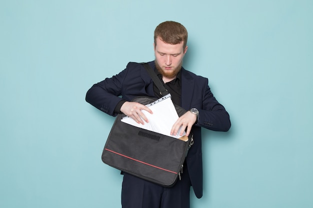 Kostenloses Foto eine junge attraktive männliche vorderansicht mit bart im schwarzen dunklen klassischen modernen anzug, der aktenrucksack auf dem blauen raum hält