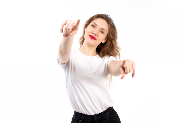 Eine junge Dame der Vorderansicht im weißen Hemd und in den schwarzen modernen Jeans, die Finger zeigen, die auf dem Weiß lächeln