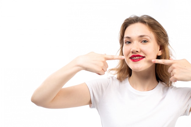 Eine junge Dame der Vorderansicht im weißen Hemd und in den schwarzen modernen Jeans, die ihre Finger zeigen, die auf dem Weiß lächeln