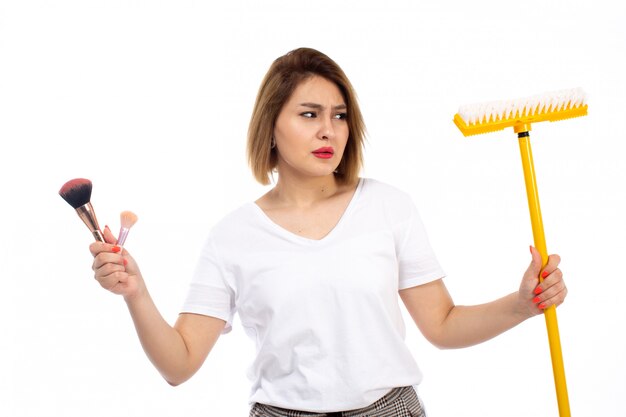 Eine junge Dame der Vorderansicht im weißen Hemd und in der hellen modernen Hose, die gelben Mopp und Make-up-Dinge auf dem Weiß hält