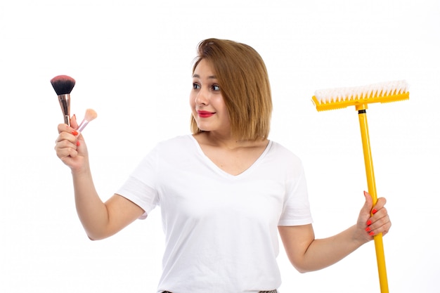 Eine junge Dame der Vorderansicht im weißen Hemd und in der hellen modernen Hose, die gelben Mopp und Make-up-Dinge auf dem Weiß hält