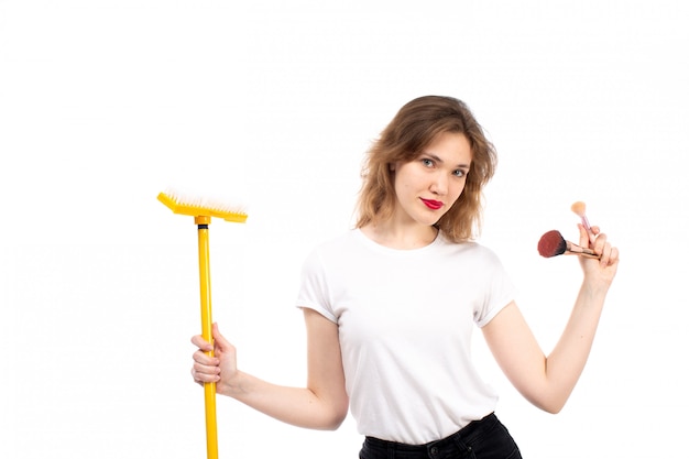 Eine junge Dame der Vorderansicht im weißen Hemd und in der schwarzen Hose, die gelben Mopp und Make-up-Dinge auf dem Weiß halten