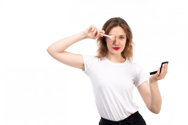 Eine junge Dame der Vorderansicht im weißen Hemd und in der schwarzen Hose, die Make-up auf dem Weiß tun