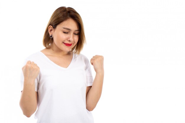 Eine junge Dame der Vorderansicht im weißen T-Shirt, das erfreut erfreut auf dem Weiß aufwirft