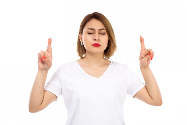 Eine junge Dame der Vorderansicht im weißen T-Shirt, das gekreuzte Finger aufwirft, schloss Augen auf dem Weiß