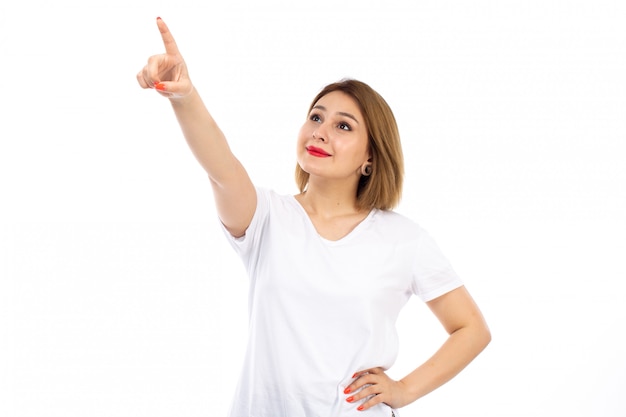 Eine junge Dame der Vorderansicht im weißen T-Shirt, das in den Himmel zeigt, der auf dem Weiß lächelt