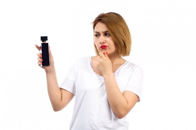 Eine junge Dame der Vorderansicht im weißen T-Shirt, das schwarze Parfümröhre hält, nicht sicherer Ausdruck auf dem Weiß