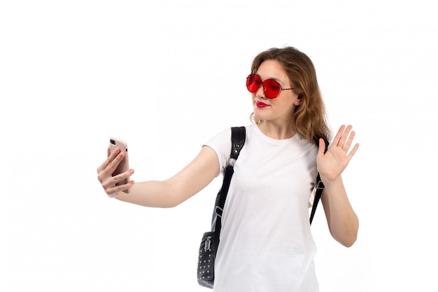 Eine junge Dame der Vorderansicht im weißen T-Shirt rote Sonnenbrille schwarze Tasche lächelnd Selfie auf dem Weiß