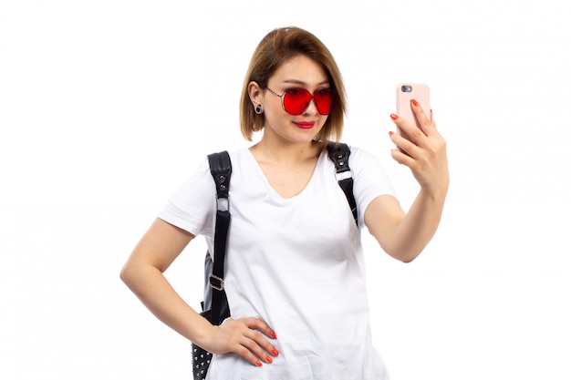 Eine junge Dame der Vorderansicht im weißen T-Shirt rote Sonnenbrille schwarze Tasche lächelnd Selfie auf dem Weiß