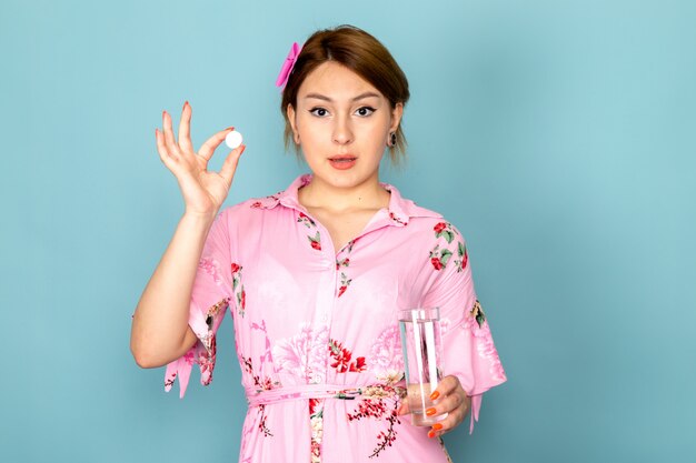 Eine junge Dame der Vorderansicht in der Blume entwarf rosa Kleid, das Pille und Glas Wasser auf Blau hält