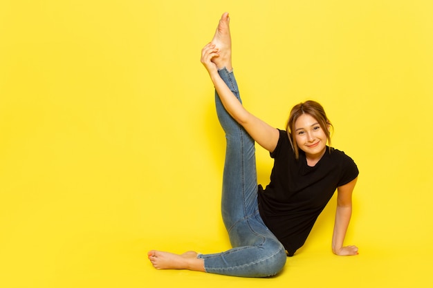 Eine junge Frau der Vorderansicht, die im schwarzen Hemd und in den blauen Jeans sitzt, die meditieren und Gymnastik auf Gelb tun