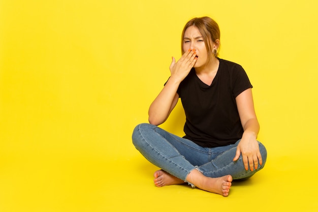 Eine junge Frau der Vorderansicht, die im schwarzen Hemd und in den blauen Jeans sitzt und auf Gelb niest