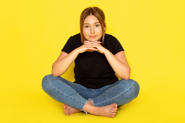 Kostenloses Foto eine junge frau der vorderansicht, die im schwarzen hemd und in den blauen jeans sitzt und mit lächeln auf gelb aufwirft