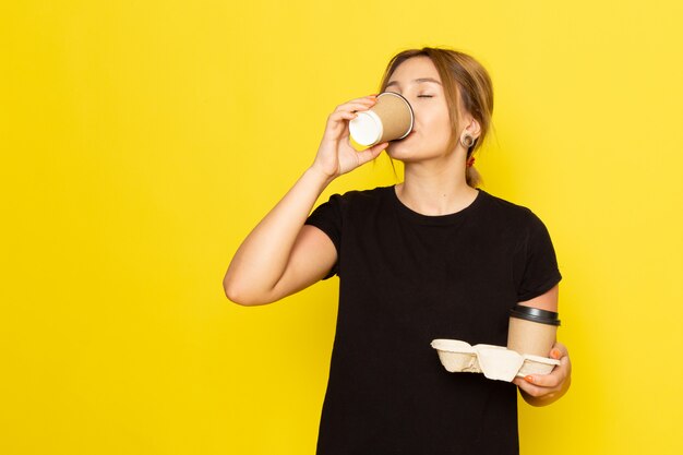 Eine junge Frau der Vorderansicht im schwarzen Kleid, die Kaffee auf Gelb trinkt