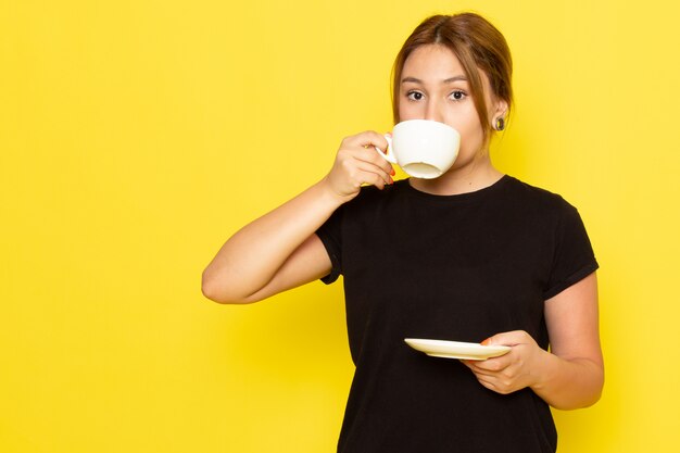Eine junge Frau der Vorderansicht im schwarzen Kleid, die Kaffee auf Gelb trinkt