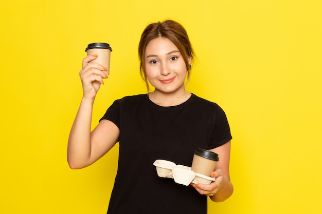 Eine junge Frau der Vorderansicht im schwarzen Kleid, die Kaffeetassen mit Lächeln auf ihrem Gesicht auf Gelb hält