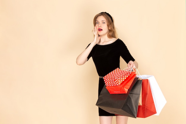 Eine junge Frau der Vorderansicht im schwarzen Kleid mit Kettengürtel, der Einkaufspakete auf Beige hält