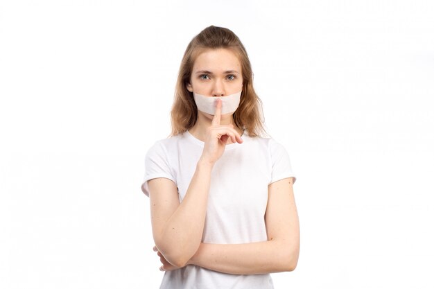 Eine junge Frau der Vorderansicht im weißen T-Shirt mit weißem Verband um ihren Mund, der aufhört, Zeichen auf dem Weiß zu sprechen