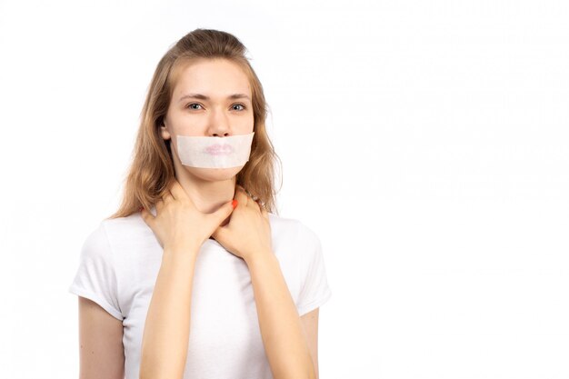 Eine junge Frau der Vorderansicht im weißen T-Shirt mit weißem Verband um ihren Mund, der physische Bedrohungen auf dem Weiß erhält