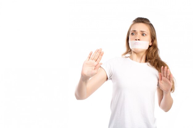 Eine junge Frau der Vorderansicht im weißen T-Shirt mit weißem Verband um ihren Mund fürchtet vorsichtig auf dem Weiß