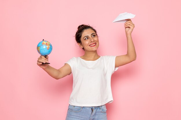 Eine junge Frau der Vorderansicht im weißen T-Shirt und in den blauen Jeans, die Papierflugzeug und kleinen Globus halten