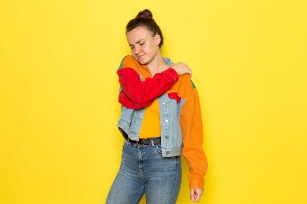 Eine junge Frau der Vorderansicht in der bunten Jacke des gelben Hemdes und der blauen Jeans, die einen Rückenschmerz haben