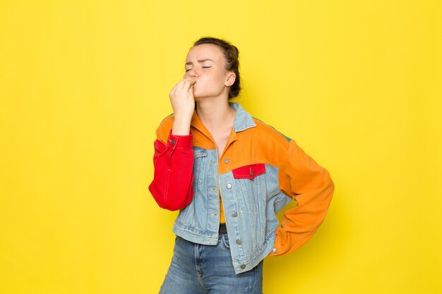 Eine junge Frau der Vorderansicht in der bunten Jacke des gelben Hemdes und der blauen Jeans, die leckeres Zeichen zeigen