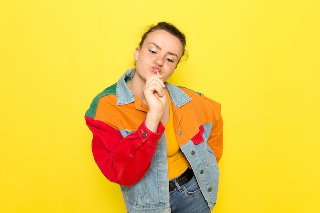 Eine junge Frau der Vorderansicht in der bunten Jacke des gelben Hemdes und im lustigen Aufstellen der blauen Jeans