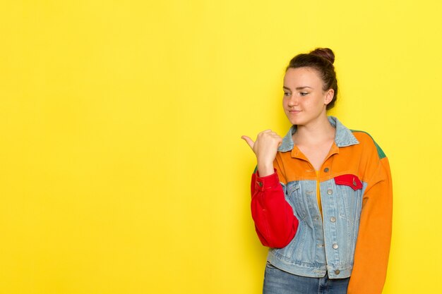 Eine junge Frau der Vorderansicht in der bunten Jacke des gelben Hemdes und in den blauen Jeans, die aufwerfen