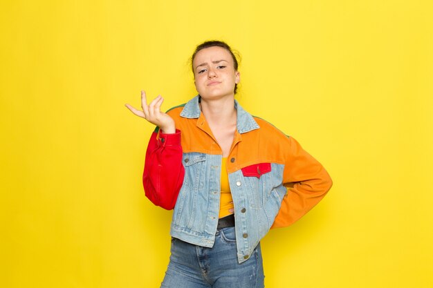 Eine junge Frau der Vorderansicht in der bunten Jacke des gelben Hemdes und in den blauen Jeans, die aufwerfen
