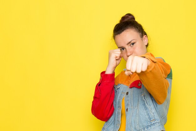 Eine junge Frau der Vorderansicht in der bunten Jacke des gelben Hemdes und in den blauen Jeans, die mit Boxstand aufwerfen