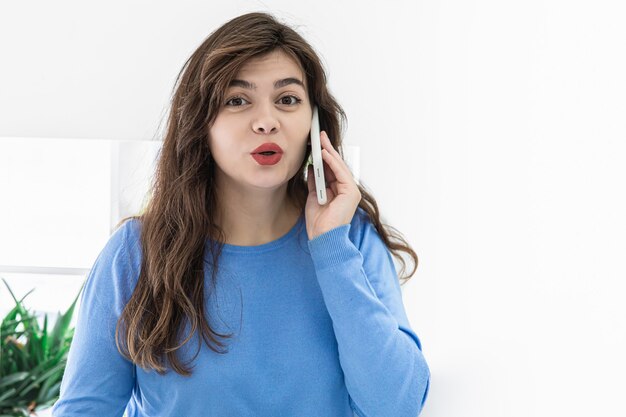 Eine junge Frau in einem blauen Pullover, brünett mit rotem Lippenstift und einem überraschten Gesicht, telefoniert in einem weißen Interieur.