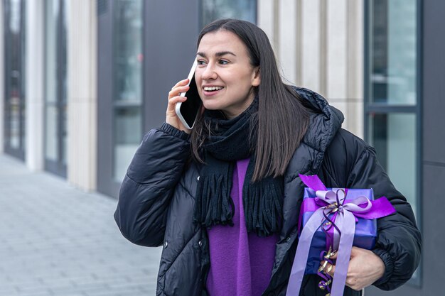 Eine junge frau mit einem lila geschenk in der hand, die draußen telefoniert