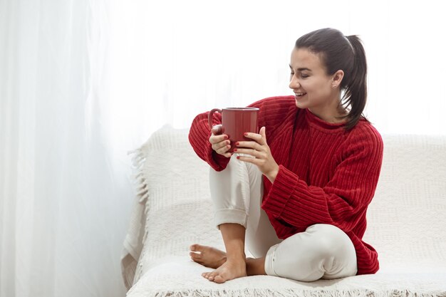 Eine junge Frau mit einer roten Tasse heißem Getränk in einem gemütlichen roten Pullover ruht zu Hause auf der Couch.