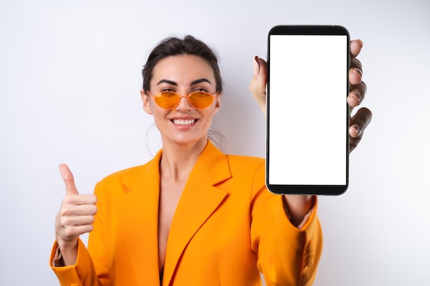 Eine junge Frau mit trendiger, stylischer Brille und einer leuchtend orangefarbenen, übergroßen Jacke auf weißem Hintergrund hält ein Telefon mit einem leeren weißen Bildschirm