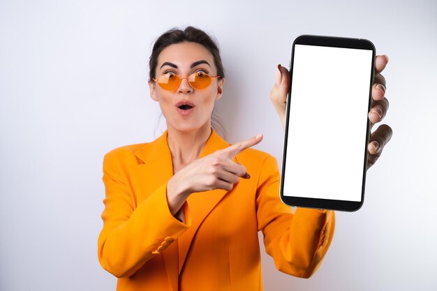Eine junge Frau mit trendiger, stylischer Brille und einer leuchtend orangefarbenen, übergroßen Jacke auf weißem Hintergrund hält ein Telefon mit einem leeren weißen Bildschirm