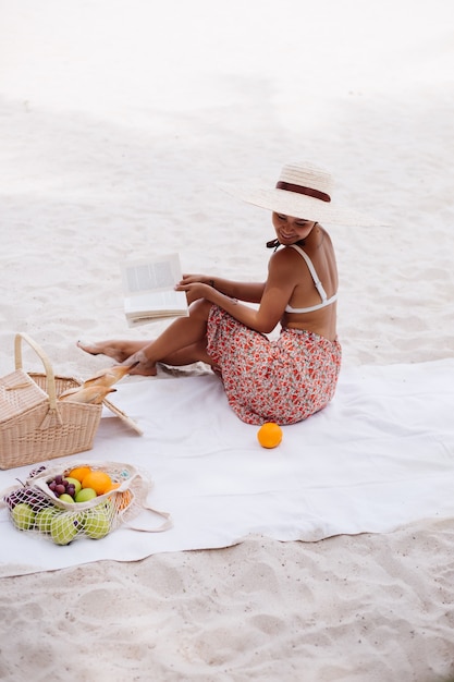 Eine junge Frau sitzt auf dem Strandteppich in einem Strohhut und einer weißen Strickkleidung