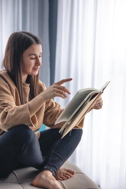 Kostenloses Foto eine junge frau sitzt auf einem sessel am fenster und liest ein buch