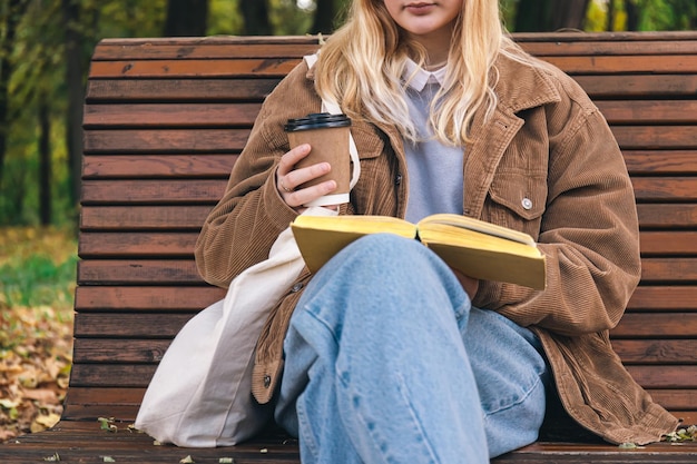 Eine junge Frau sitzt auf einer Bank in einem Herbstpark, trinkt Kaffee und liest ein Buch